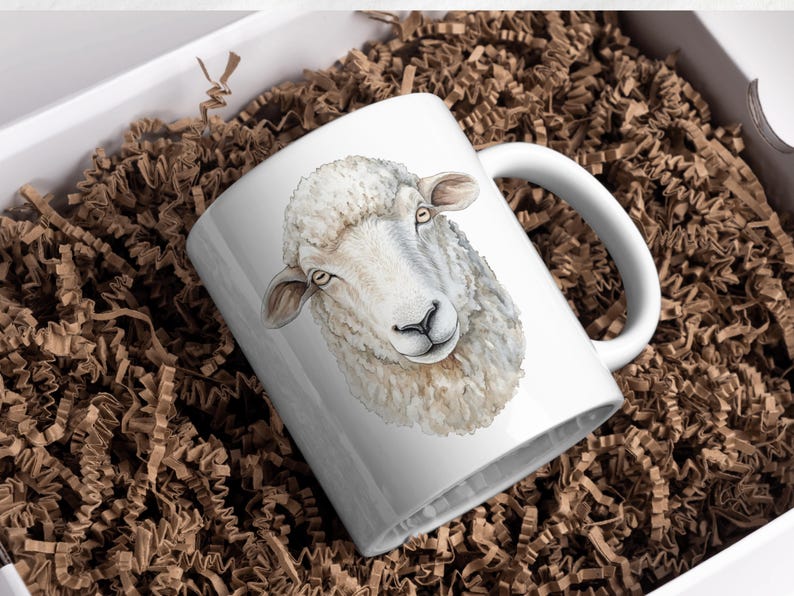May include: White ceramic mug featuring a watercolor illustration of a sheep's head. The sheep has fluffy white wool, brown ears, and a black nose. The mug is presented in a white box with brown paper filler.