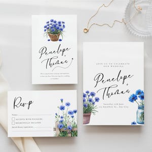 May include: A set of wedding stationery with a floral design. The invitation, RSVP card, and details card feature blue watercolour flowers and the names "Penelope + Thomas". The suite has a rustic, floral theme.