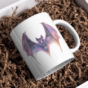 Pu&ograve; includere: Tazza in ceramica bianca con un'illustrazione ad acquerello di un pipistrello con ali viola e arancioni. Il pipistrello &egrave; centrato sulla tazza. La tazza &egrave; in una scatola bianca con imbottitura di carta marrone.