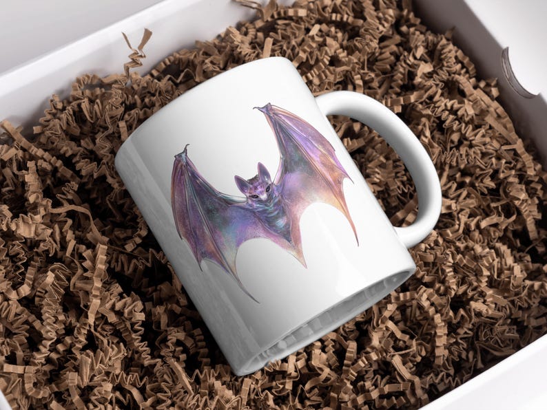 Pu&ograve; includere: Tazza in ceramica bianca con un'illustrazione ad acquerello di un pipistrello con ali viola e arancioni. Il pipistrello &egrave; centrato sulla tazza. La tazza &egrave; in una scatola bianca con imbottitura di carta marrone.