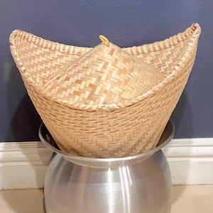 May include: A traditional Thai sticky rice steamer set consisting of a woven bamboo conical basket with a small yellow loop at the top, resting securely inside a polished silver-colored aluminum pot with a wide, flared rim and a rounded base.