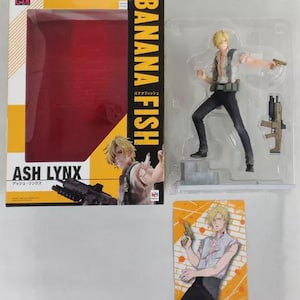 May include: A Banana Fish Ash Lynx collectible figure set including a display box with yellow and black branding, a plastic-encased blonde character figure in a white shirt and black pants holding a handgun, and a rectangular character card.