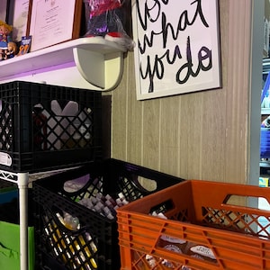 May include: A white shelf with two framed diplomas and a stuffed bear in a plastic bag. The shelf is mounted on a wall with wood paneling. A framed sign with the text "Love what you do" is hanging on the wall. Below the shelf are three black plastic crates stacked on top of each other. Next to the crates is a red plastic crate.