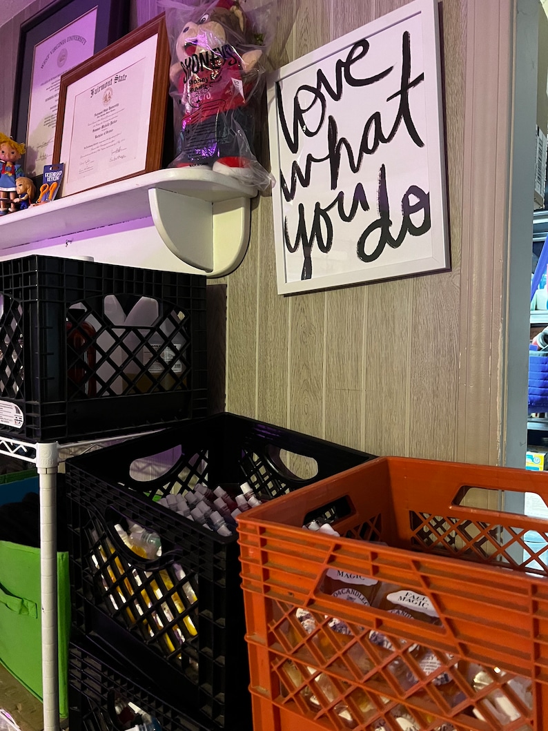 May include: A white shelf with two framed diplomas and a stuffed bear in a plastic bag. The shelf is mounted on a wall with wood paneling. A framed sign with the text "Love what you do" is hanging on the wall. Below the shelf are three black plastic crates stacked on top of each other. Next to the crates is a red plastic crate.
