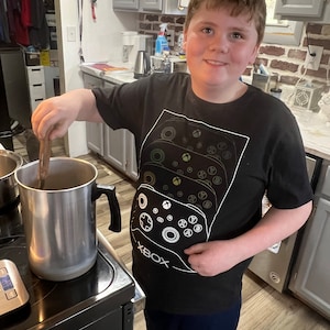 May include: A young person wearing a black t-shirt with a white Xbox controller graphic and the word "XBOX" printed on the front. The person is standing in a kitchen and stirring a pot on the stove.