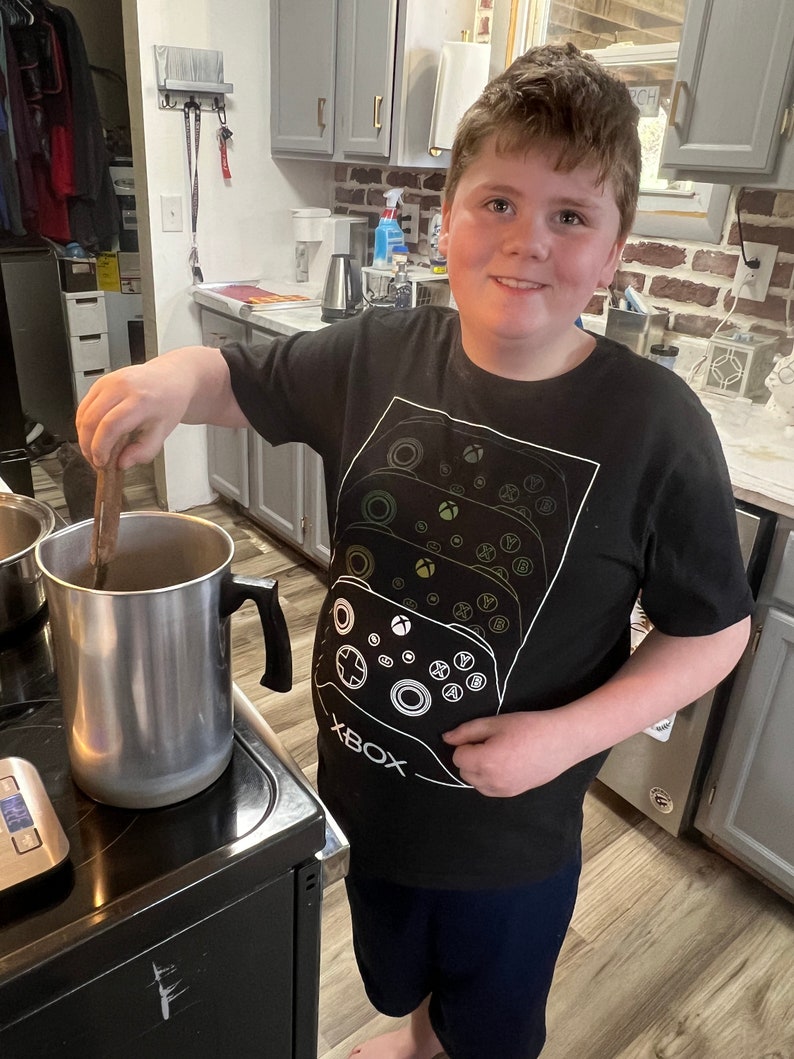 May include: A young person wearing a black t-shirt with a white Xbox controller graphic and the word "XBOX" printed on the front. The person is standing in a kitchen and stirring a pot on the stove.