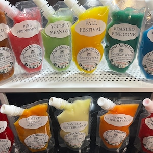 May include: A collection of ten colorful pouches filled with wax melts. Each pouch has a different scent and a label with the name of the scent. The scents include Marshmallow Cocoa, Pink Peppermint, You're a Mean One, Fall Festival, Roasted Pine Cone, Blueberry Cobbler, Apple Cinnamon, Pumpkin Pecan Waffles, Vanilla Buttercream, and Pumpkin Marshmallow. The pouches are all labeled with the company name "304CANDLECO" and the words "SQUEEZY WAX".