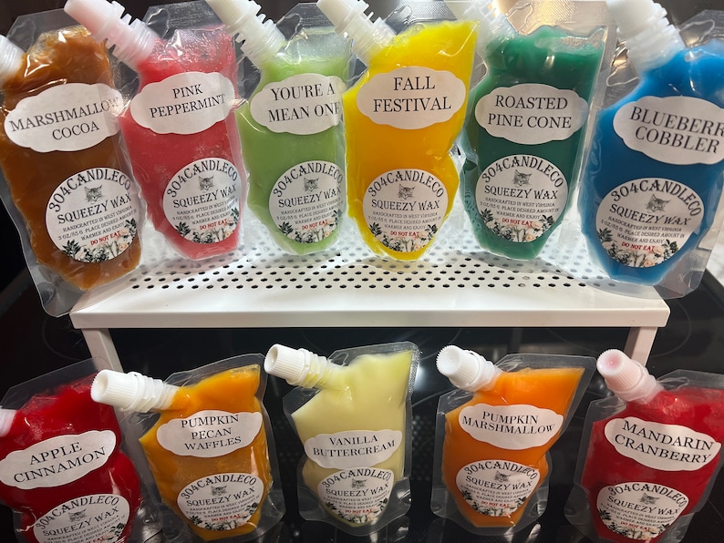 May include: A collection of ten colorful pouches filled with wax melts. Each pouch has a different scent and a label with the name of the scent. The scents include Marshmallow Cocoa, Pink Peppermint, You're a Mean One, Fall Festival, Roasted Pine Cone, Blueberry Cobbler, Apple Cinnamon, Pumpkin Pecan Waffles, Vanilla Buttercream, and Pumpkin Marshmallow. The pouches are all labeled with the company name "304CANDLECO" and the words "SQUEEZY WAX".