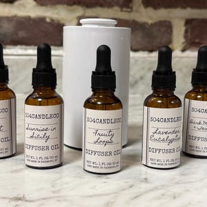 May include: Five amber glass diffuser oil bottles with black droppers and white labels. The labels read "Aruba Coconut", "Sunrise in Sicily", "Fruity Loops", "Lavender Eucalyptus", and "Dark Mahogany & Teakwood". A white ceramic vase is in the background.