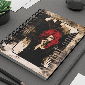 May include: A spiral bound notebook featuring a watercolour illustration of a woman with red hair sitting with a black bird on her shoulder. The background is a cityscape with a dark, moody feel.