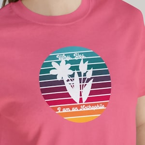 May include: A pink t-shirt featuring a circular graphic with a floral design and the text "Why, Yes, I am an Anthophile." The graphic has a retro sunset design with horizontal stripes in various colors.