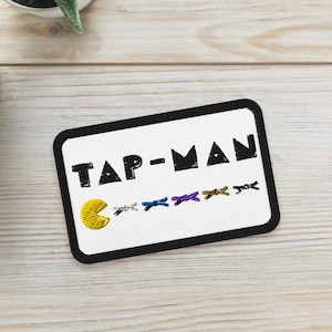 以下が含まれることがあります： 黒い文字で「TAP-MAN」と書かれた白い長方形のパッチ。黄色いパックマンのキャラクターとカラフルなゴーストが特徴です。明るい木目調の背景に、小さな鉢植えの多肉植物が2つあります。