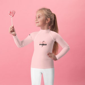May include: A light pink long-sleeved shirt with the text "GIRL POWER" and a lightning bolt graphic. The shirt is paired with white pants. The background is a solid pink color.