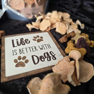 May include: A wooden sign with the text "Life is Better With Dogs" and paw prints, surrounded by dog treats. The treats are various shapes and colours, including light brown and purple. A bag of sliced treats is in the background.