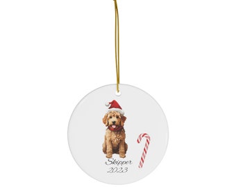 May include: White ceramic Christmas ornament featuring a golden doodle wearing a Santa hat and a red collar. A candy cane and the name "Skipper 2023" are also on the ornament. A gold string is attached for hanging.