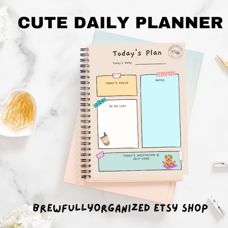 May include: A daily planner with a light brown cover and a spiral binding. The cover has a cute illustration of a bubble tea cup and a dog meditating. The planner has sections for today's date, today's focus, to do list, notes, and today's meditation and self-care.