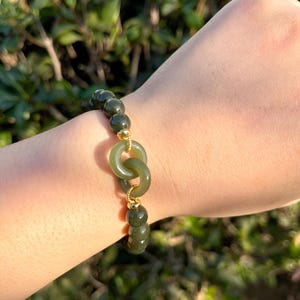May include: A green jade bead bracelet with a gold double ring charm. The bracelet is on a person's wrist.