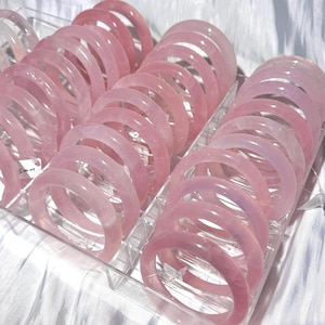 May include: A collection of pink rose quartz bracelets displayed in a clear plastic organizer. The bracelets are a soft pink color and appear smooth and polished. The bracelets are round and have a consistent width. The background is a white, textured surface.