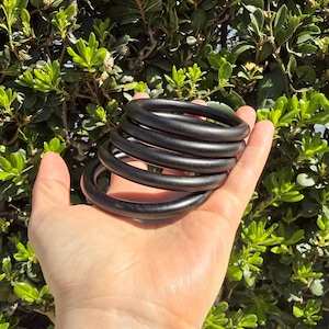 May include: A hand holds a stack of six black wooden bangles. The bangles are smooth and round, with a dark, matte finish. The background is a lush green bush.