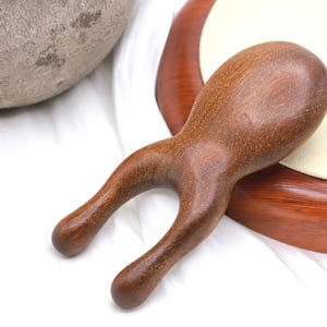 May include: A wooden massage tool with a curved handle and two rounded prongs. The tool is a warm brown color and appears to be made of polished wood. It is resting on a white surface with a wooden tray and a gray stone pot in the background.