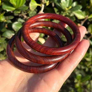 May include: Three wooden bangles in a deep red hue with a natural wood grain pattern. The bangles are stacked on top of each other.