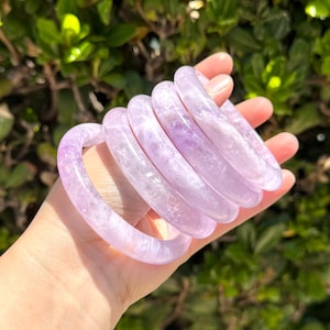 May include: A stack of five smooth, translucent light purple amethyst bangle bracelets. Each circular bracelet features a polished finish with natural crystalline inclusions visible within the stone, creating a soft lavender hue throughout the set.