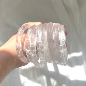 May include: A collection of clear quartz crystal bangles. The bracelets are round and transparent, with visible internal textures. The light catches the crystal, creating highlights and shadows. The bangles are stacked together.