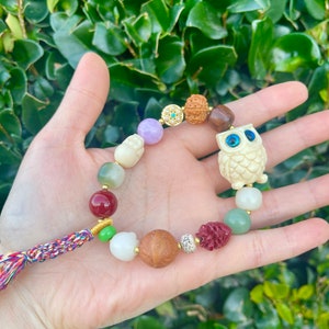 May include: A white owl charm bracelet with colorful beads, including red, green, brown, and purple. The bracelet has a tassel made of multi-colored threads.
