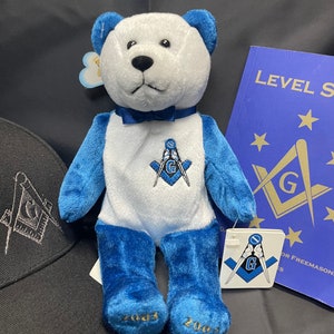 May include: A blue and white teddy bear wearing a bow tie and a Masonic symbol on its chest. The bear is sitting on a black surface with a black baseball cap and a blue book with the text "LEVELS" on the cover.