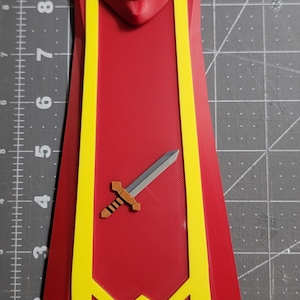 May include: A red and yellow 3D-printed cape with a sword emblem. The cape features a hood and yellow trim, with a sword design on the front. The item is likely a costume piece or a decorative item.