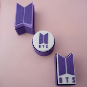 May include: Three purple and white BTS logo stamps on a pink surface. The stamps are in different shapes, including a circle and two rectangular shapes. The BTS logo is visible on each stamp.