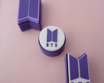 May include: Three purple and white BTS logo stamps on a pink surface. The stamps are in different shapes, including a circle and two rectangular shapes. The BTS logo is visible on each stamp.