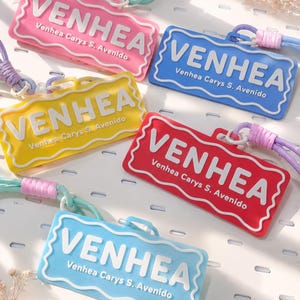 Op de afbeelding: Een verzameling kleurrijke rechthoekige labels in roze, blauw, geel, rood en lichtblauw. Elk label toont de naam "VENHEA" in wit, samen met de tekst "Venhea Carys S. Avenido". De labels hebben een golvende rand en zijn bevestigd aan een koord.