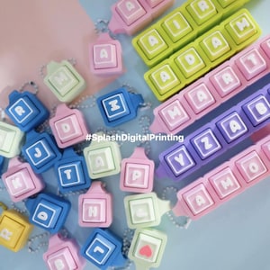 Könnte beinhalten: Eine Sammlung farbenfroher Schlüsselanhänger und Zubehörteile in Form von Tastenkappen. Die Schlüsselanhänger sind in verschiedenen Farben erhältlich, darunter Rosa, Blau, Grün und Lila, mit weißen Buchstaben und Symbolen. Der Text "#SplashDigitalPrinting" ist sichtbar.