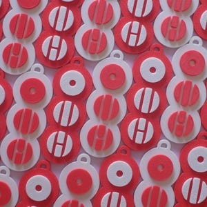 May include: A close-up of multiple red, white, and red-and-white layered ornaments. Each ornament has a small loop at the top. Some have the letter "H" in the centre, while others have a circle or stripes.