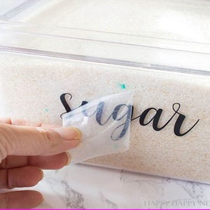 K&ouml;nnte beinhalten: Eine Hand zieht eine transparente Plastikfolie von einem durchsichtigen Plastikbeh&auml;lter mit wei&szlig;em Zucker ab. Der Beh&auml;lter ist mit "Sugar" in schwarzer Kursivschrift beschriftet.