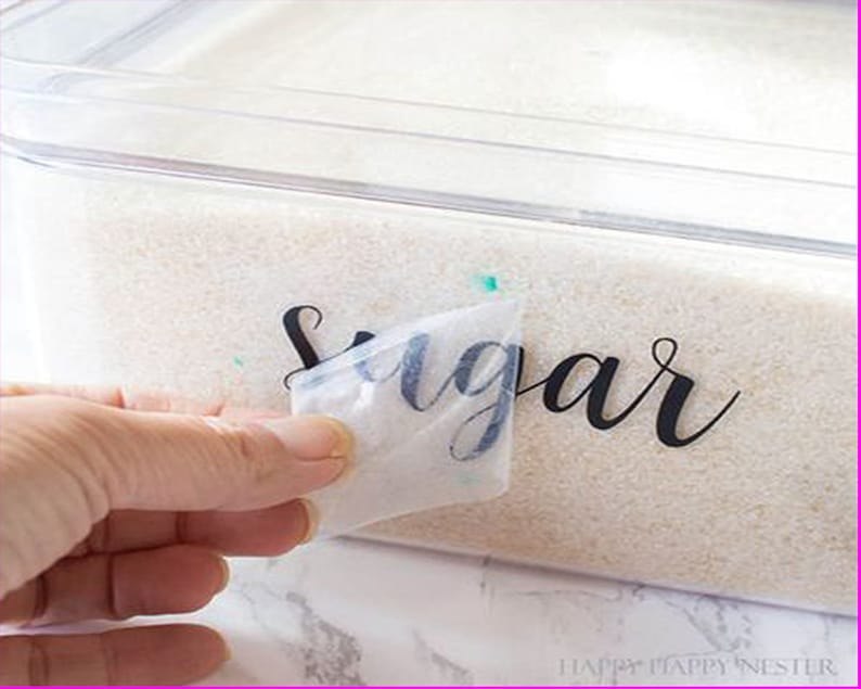 K&ouml;nnte beinhalten: Eine Hand zieht eine transparente Plastikfolie von einem durchsichtigen Plastikbeh&auml;lter mit wei&szlig;em Zucker ab. Der Beh&auml;lter ist mit "Sugar" in schwarzer Kursivschrift beschriftet.