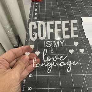 K&ouml;nnte beinhalten: Wei&szlig;er Vinyl-Aufkleber mit dem Text "Coffee is my love language" mit kleinen Herzen um den Text. Der Aufkleber ist auf einem transparenten Hintergrund.