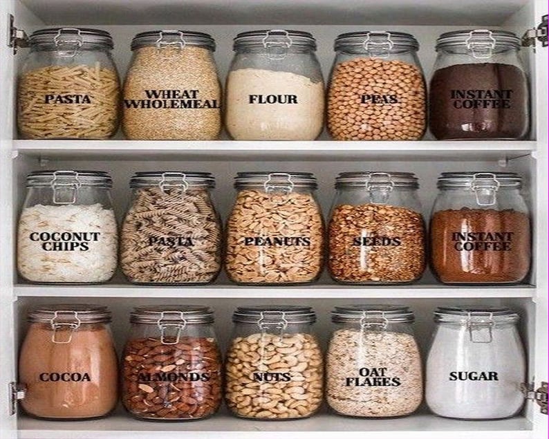 K&ouml;nnte beinhalten: Ein Satz von 15 Glasbeh&auml;ltern mit Etiketten f&uuml;r verschiedene Vorratsprodukte, darunter Pasta, Weizen, Mehl, Erbsen, Instantkaffee, Kokoschips, Erdn&uuml;sse, Samen, Kakao, Mandeln, N&uuml;sse, Haferflocken und Zucker. Die Beh&auml;lter sind in drei Reihen auf einem wei&szlig;en Regal angeordnet.