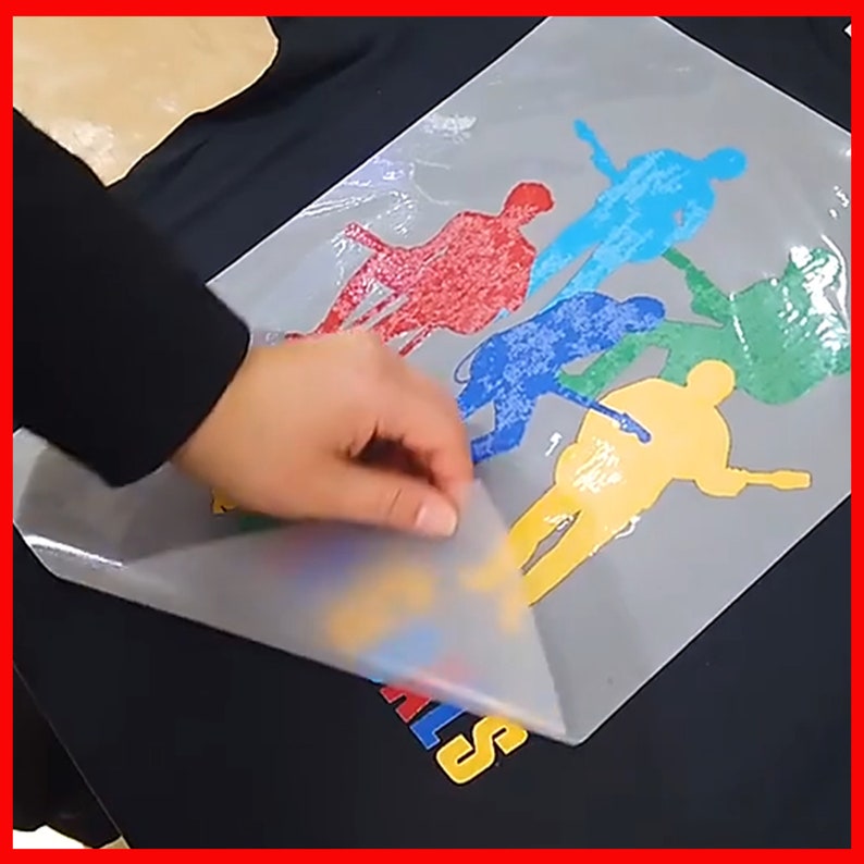May include: A hand peeling back a clear sheet of transfer paper revealing a colorful design of five figures on a black t-shirt. The figures are silhouetted and are in shades of red, blue, green, and yellow.