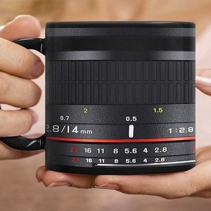 K&ouml;nnte beinhalten: Schwarze Keramiktasse in Form eines Kameraobjektivs mit dem Text "2.8/14 mm" und "1:2.8" auf der Seite. Die Tasse hat einen roten Ring um den Boden und einen schwarzen Griff.