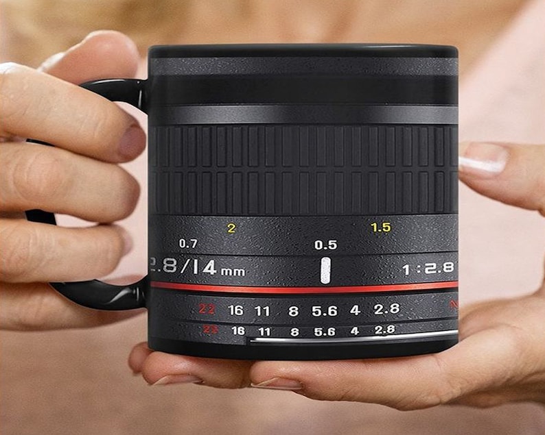 K&ouml;nnte beinhalten: Schwarze Keramiktasse in Form eines Kameraobjektivs mit dem Text "2.8/14 mm" und "1:2.8" auf der Seite. Die Tasse hat einen roten Ring um den Boden und einen schwarzen Griff.