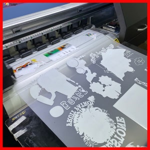 May include: A close-up of a large format printer printing white vinyl designs on a gray background. The designs include a silhouette of a person, a flower, and the word "DUKES".