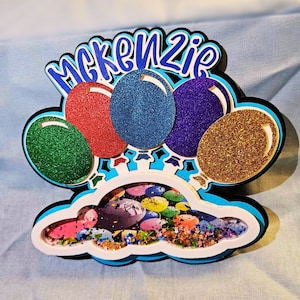 May include: A blue and white cloud-shaped frame with a glitter-filled center. Five balloons in red, green, blue, purple, and gold are attached to the top of the frame. The word "MCKENZIE" is written in black letters on the top of the frame.
