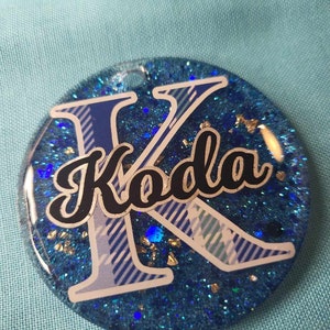 May include: A round, blue glitter keychain with the name "Koda" in black script. The letter "K" is in a blue and white plaid pattern. The keychain has a small hole at the top for attachment.