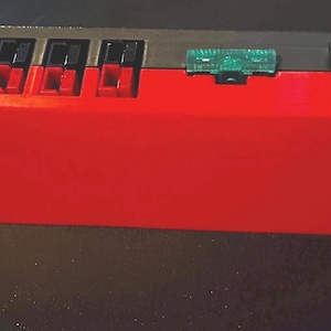 Puede incluir: Un dispositivo electrónico rojo y negro con múltiples interruptores negros y un componente verde. El dispositivo tiene un interruptor de encendido/apagado negro y es de forma rectangular.