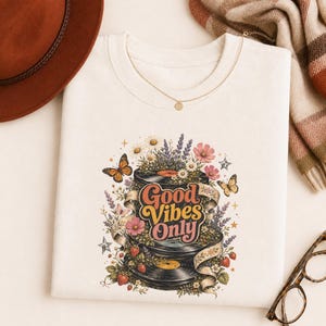 May include: A cream-colored t-shirt featuring a graphic print of two stacked black vinyl records wrapped in a ribbon with the text Good Vibes Only. The design includes colorful illustrations of daisies, lavender, pink flowers, strawberries, and butterflies.