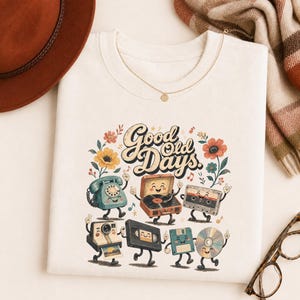 Könnte beinhalten: Ein cremefarbenes T-Shirt mit einem Grafikdruck und dem Text Good Old Days. Das Design zeigt anthropomorphe Illustrationen eines Wählscheibentelefons, eines Plattenspielers, einer Kassette, einer Kamera, einer VHS-Kassette, einer Diskette und einer CD.