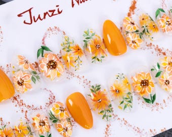 May include: A set of ten false nails with a clear base and a floral design featuring yellow and orange flowers. The nails are displayed on a white surface with the text "Junzi Nail" written in red.