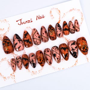 May include: Set of 14 Halloween-themed false nails with a variety of designs, including pumpkins, bats, and cobwebs. The nails are a light peach color with black and orange accents. Junzi Nail is written in red at the top of the image.